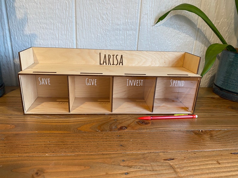 May include: A wooden money organizer with four compartments labeled "SAVE", "GIVE", "INVEST", and "SPEND". The name "LARISA" is above the compartments. A red pen rests in front of the organizer. The organizer is made of light-colored wood.