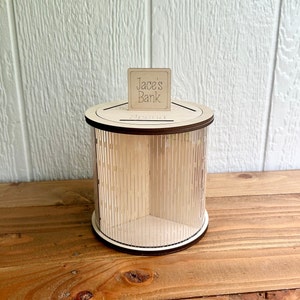 May include: Wooden coin bank with a clear acrylic cylinder and a slot on the top. The bank has a sign that says "Jace's Bank" and the word "Spend" is written on the top of the bank.