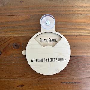 May include: A light-colored wooden office sign with the text "PLEASE KNOCK" above "WELCOME TO KELLY'S OFFICE". The sign has a circular shape with a small handle and a clear suction cup for mounting.