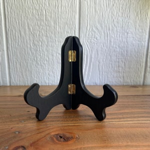May include: A black wooden easel stand with gold hinges. The stand is designed to hold a picture or other object upright.