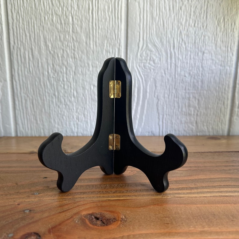May include: A black wooden easel stand with gold hinges. The stand is designed to hold a picture or other object upright.