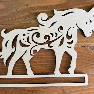 May include: White wooden unicorn cutout with intricate floral designs. The unicorn has a long, flowing mane and tail. The cutout is attached to a wooden base.