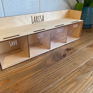May include: A wooden money organizer with four compartments labeled "SAVE", "GIVE", "INVEST", and "SPEND". The name "LARISA" is laser-cut on the top. The organizer is made of light-colored wood and has clear dividers.