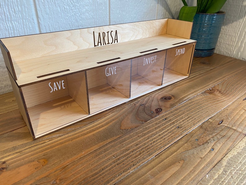May include: A wooden money organizer with four compartments labeled "SAVE", "GIVE", "INVEST", and "SPEND". The name "LARISA" is laser-cut on the top. The organizer is made of light-colored wood and has clear dividers.
