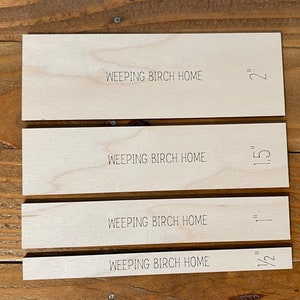 May include: Four rectangular wooden pieces with the text "WEEPING BIRCH HOME" and measurements of 2", 1.5", 1", and 0.5" etched on them. The pieces are arranged on a wooden surface.