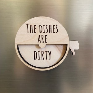May include: A wooden round sign with the text "The Dishes Are Dirty" and a thumbs down icon. The sign is attached to a refrigerator with a magnet.