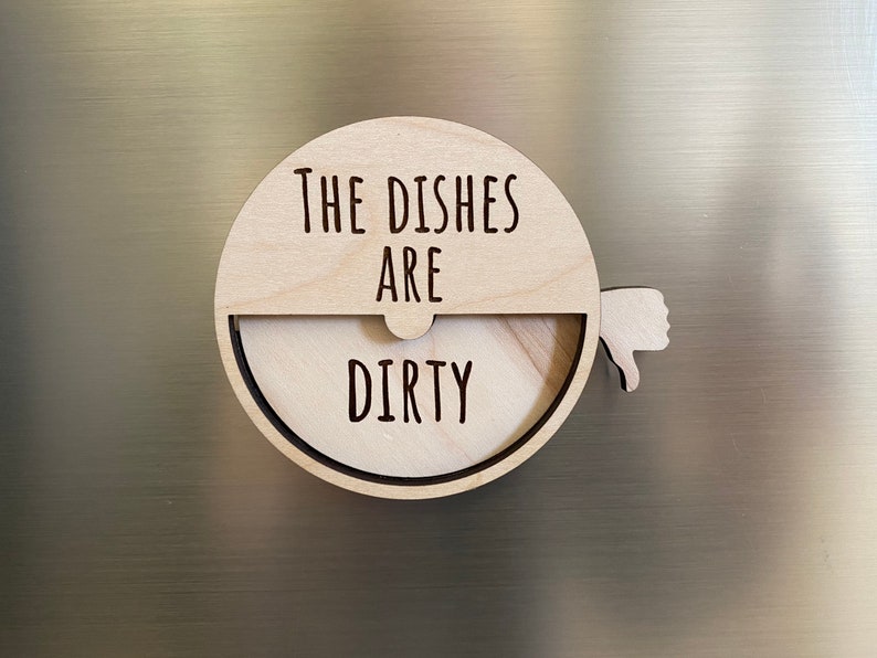 May include: A wooden round sign with the text "The Dishes Are Dirty" and a thumbs down icon. The sign is attached to a refrigerator with a magnet.