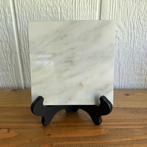 May include: A white marble tile with a light gray veining pattern, displayed on a black easel stand.