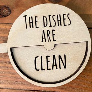 May include: A wooden sign with a sliding panel that reads "The Dishes Are Clean". The sign is made of light wood and has a black border.