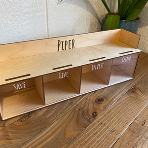 May include: A wooden money organizer with four compartments labeled "SAVE", "GIVE", "INVEST", and "SPEND". The name "PIPER" is above the compartments. The organizer is made of light-colored wood and sits on a wooden surface.