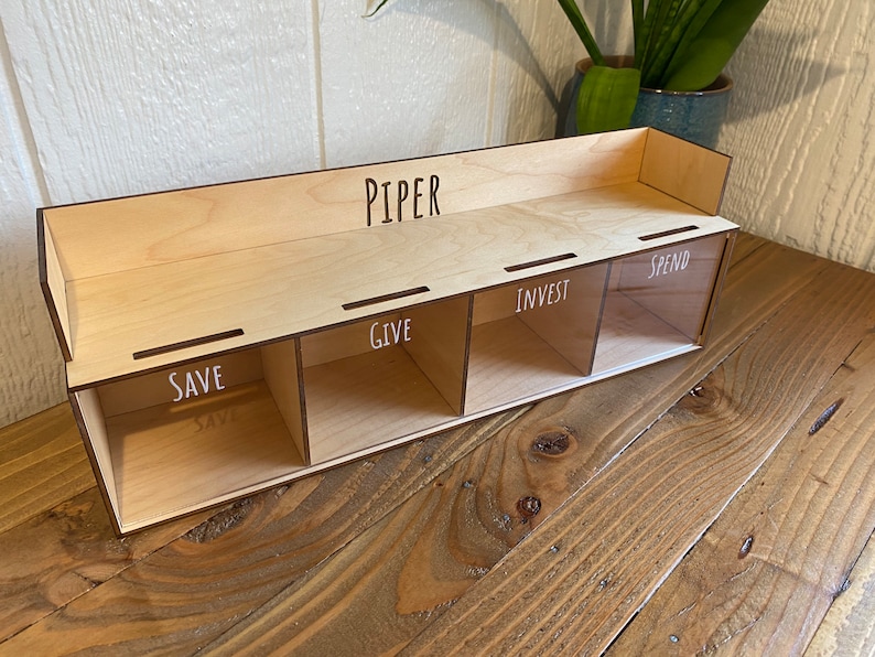 May include: A wooden money organizer with four compartments labeled "SAVE", "GIVE", "INVEST", and "SPEND". The name "PIPER" is above the compartments. The organizer is made of light-colored wood and sits on a wooden surface.