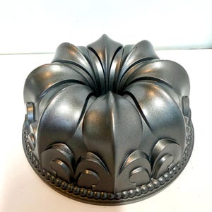 May include: A metallic dark gray bundt cake pan featuring a deep, sculpted floral or petal-like design radiating from the center. The base of the pan is encircled by a decorative beaded rim, and the surface has a smooth, reflective finish.