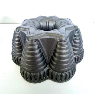 May include: A dark gray metallic bundt cake pan featuring a complex, multi-faceted geometric design with deep, ribbed vertical fluting and a central hollow tube. The exterior surface has a smooth, matte finish with distinct angular structural ridges.