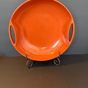 May include: A round, bright orange ceramic serving dish with two integrated side handles. The dish features a smooth, glossy finish with subtle tonal variations in the orange glaze, and it is displayed on a dark metal wire stand.