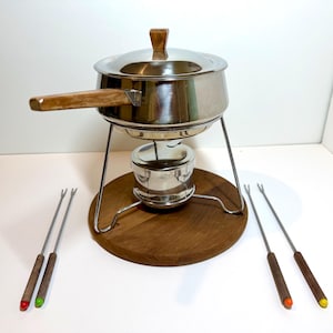 May include: A stainless steel fondue pot with a lid, featuring a wooden handle and knob. The pot rests on a metal stand over a burner, all set on a circular wooden base. Four metal fondue forks with wooden handles and colored tips are included.