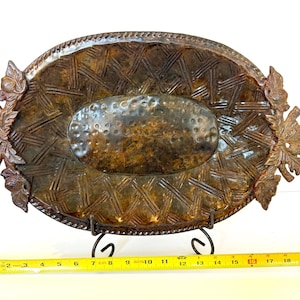 May include: An oval-shaped brown decorative tray with a woven basket-weave pattern and a hammered center. The edges feature sculpted acorn and leaf motifs. A yellow measuring tape placed in front shows the tray is approximately 19.5 inches wide.