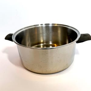 May include: A round, stainless steel cooking pot with a polished metallic finish. The pot features two black, heat-resistant side handles attached to the exterior rim. The interior is smooth and reflective, showing a deep, open basin design.
