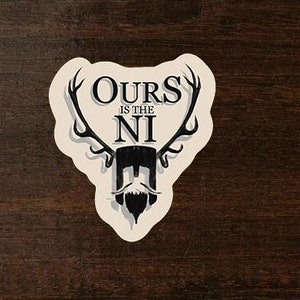 May include: A cream-colored sticker with the text "OURS IS THE NI" in a stylized font, featuring antlers and a beard design. The sticker is on a dark wood surface, with a partial view of a coffee cup in the upper right corner.