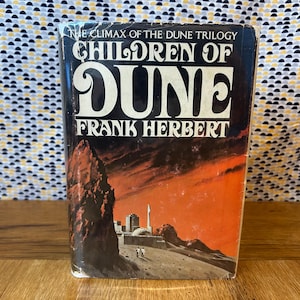 May include: A vintage paperback book titled "Children of Dune" by Frank Herbert. The cover features a desert landscape with a city and figures, set against a fiery orange and red sky. The title is in large, bold white letters.