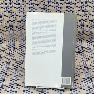 May include: The back cover of a book titled "The Economics of Feasible Socialism" by Alec Nove. The book cover is gray with white text. The text describes the book as a readable, controversial, and insightful analysis of the Soviet economic system.