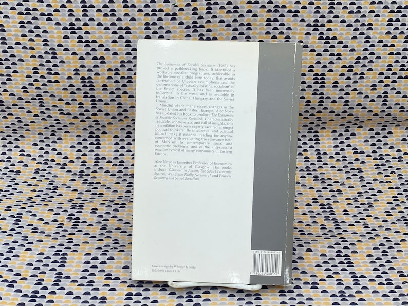 May include: The back cover of a book titled "The Economics of Feasible Socialism" by Alec Nove. The book cover is gray with white text. The text describes the book as a readable, controversial, and insightful analysis of the Soviet economic system.