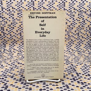 Puede incluir: Libro de bolsillo vintage titulado "The Presentation of Self in Everyday Life" de Erving Goffman. El libro tiene una cubierta color crema con texto negro y est&aacute; apoyado en un soporte de metal blanco. El fondo es una tela estampada.