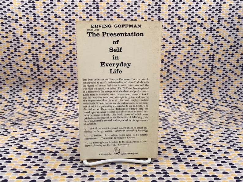 Puede incluir: Libro de bolsillo vintage titulado "The Presentation of Self in Everyday Life" de Erving Goffman. El libro tiene una cubierta color crema con texto negro y est&aacute; apoyado en un soporte de metal blanco. El fondo es una tela estampada.