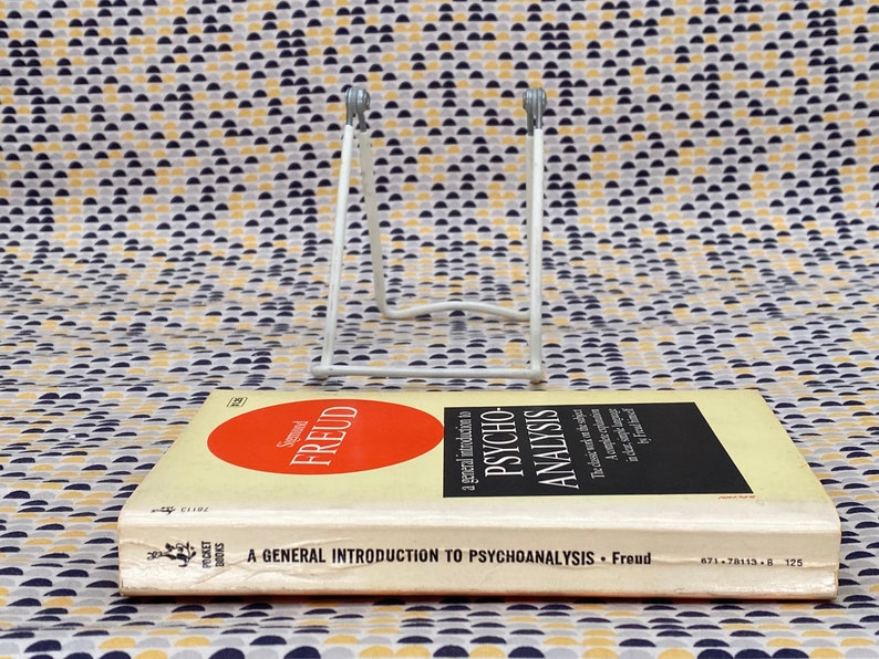 May include: A white metal book stand with a book titled "A General Introduction to Psychoanalysis - Freud" on top of it. The book cover is yellow with a red circle that says "Sigmund Freud" in white text.