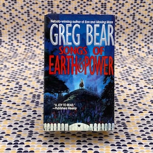 May include: A book cover with a dark blue background and a white title that reads "Songs of Earth & Power" by Greg Bear. The author is described as a Nebula-winning author of Eon and Moving Mars. The cover features a dark forest scene with a figure on horseback in the distance.