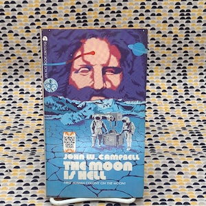 May include: A blue and white paperback book with the title "The Moon is Hell" by John W. Campbell. The cover features a man with a beard and a futuristic scene with two astronauts.