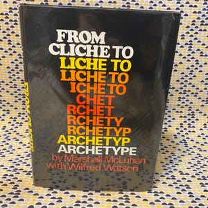 May include: A black book titled "FROM CLICHE TO ARCHETYPE" by Marshall McLuhan with Wilfred Watson. The title is in white, yellow, orange, and red lettering. The book is on a patterned background.