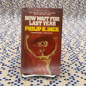 Puede incluir: Un libro de bolsillo vintage titulado "Now Wait for Last Year" de Philip K. Dick. La portada presenta una ilustración surrealista de una figura con los brazos levantados, un ojo y el texto "Compelling reading - Library Journal."