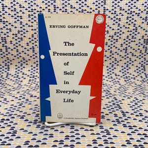 Puede incluir: Un libro de bolsillo vintage titulado "The Presentation of Self in Everyday Life" de Erving Goffman. La portada del libro presenta un fondo blanco con formas geom&eacute;tricas azules y rojas. El libro est&aacute; apoyado en un soporte de metal blanco.