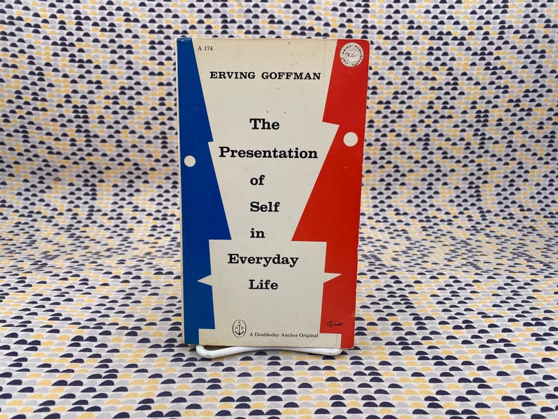 Puede incluir: Un libro de bolsillo vintage titulado "The Presentation of Self in Everyday Life" de Erving Goffman. La portada del libro presenta un fondo blanco con formas geom&eacute;tricas azules y rojas. El libro est&aacute; apoyado en un soporte de metal blanco.