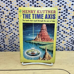 Op de afbeelding: Een vintage paperback boek getiteld "The Time Axis" van Henry Kuttner. De cover toont een kleurrijke illustratie van een landschap met een groot standbeeld, een vliegende schotel en de tekst "The battle that raged through the core of time."