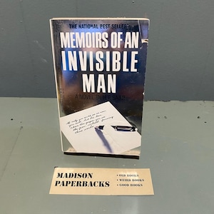 Könnte beinhalten: Ein Taschenbuch mit dem Titel Memoirs of an Invisible Man, ein Roman von H.F. Saint, mit dunkelblauem Einband, weißem Text und einer Abbildung eines Stifts, der auf Papier schreibt. Unter dem Buch liegt eine rechteckige Karte Madison Paperbacks.