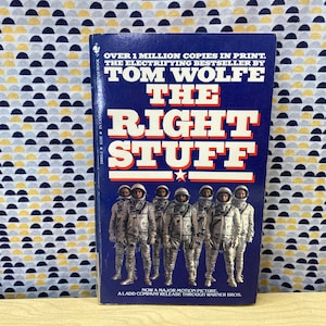 Puede incluir: Una portada de libro de bolsillo con fondo azul y texto blanco que dice "OVER 1 MILLION COPIES IN PRINT. THE ELECTRIFYING BESTSELLER BY TOM WOLFE THE RIGHT STUFF NOW A MAJOR MOTION PICTURE A LADD COMPANY RELEASE THROUGH WARNER BROS.". La portada presenta un grupo de siete astronautas con trajes espaciales blancos de pie en fila.