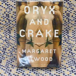 May include: A book cover with a painting of two nude figures holding fruit. The title of the book is "Oryx and Crake" by Margaret Atwood. The book is a novel.