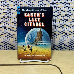 Op de afbeelding: Een vintage paperback boek getiteld "Earth's Last Citadel" met een sciencefiction illustratie. De cover toont een kasteel op een maan, figuren op een planeet en de namen van de auteurs. Het boek wordt gepresenteerd op een witte standaard.