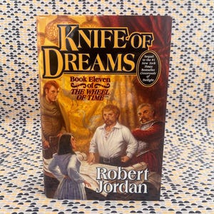 May include: A hardcover book titled "Knife of Dreams" by Robert Jordan. The cover features illustrations of several characters and the text "Book Eleven of The Wheel of Time". The book is a sequel to the #1 New York Times Bestseller "Crossroads of Twilight".