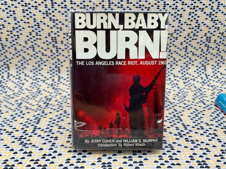 May include: A vintage paperback book titled "Burn, Baby Burn! The Los Angeles Race Riot, August 1965" by Jerry Cohen and William S. Murphy. The cover features a black and white photo of a man in a military uniform holding a rifle, with other figures in the background.