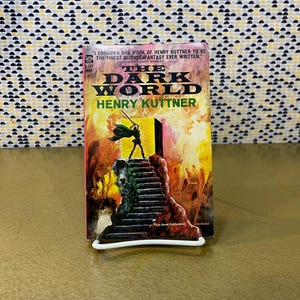 Op de afbeelding: Een paperback boek getiteld "The Dark World" van Henry Kuttner, met een levendige omslagillustratie. De omslag toont een figuur met een zwaard op een trap tegen een vurige achtergrond. Het boek staat op een witte standaard.