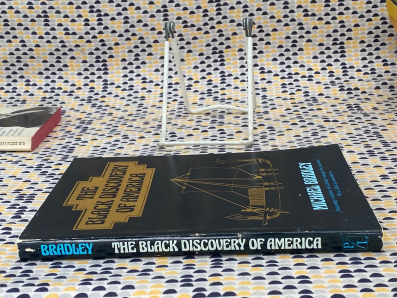 May include: A black book titled "The Black Discovery of America" by Michael Bradley. The book's cover features gold lettering and an illustration of a ship. The book is displayed on a white metal stand against a patterned background.