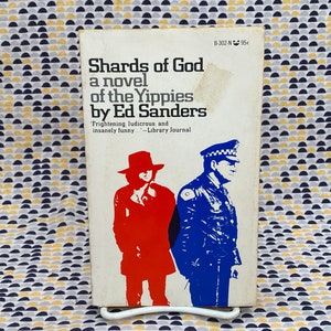 Puede incluir: Una portada de libro de bolsillo vintage con fondo blanco. El título es "Shards of God: A Novel of the Yippies" de Ed Sanders. La portada presenta una ilustración en blanco y negro de un hombre con uniforme de policía y un hombre con chaqueta y sombrero rojos.