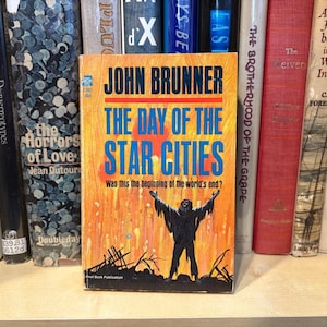 Puede incluir: Un libro de bolsillo vintage titulado "The Day of the Star Cities" de John Brunner. La portada presenta la silueta de una persona con los brazos levantados sobre un fondo naranja y amarillo. El libro está entre otros libros en una estantería.