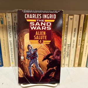 May include: A paperback book titled "The Sand Wars: Alien Salute 4" by Charles Ingrid. The cover art depicts a futuristic soldier with a weapon in a red and brown alien landscape. Other books are visible in the background.
