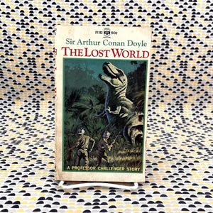 May include: Vintage paperback book titled "The Lost World" by Sir Arthur Conan Doyle. The cover features a green and white illustration of a dinosaur and explorers in a jungle setting. The book is set against a patterned background.