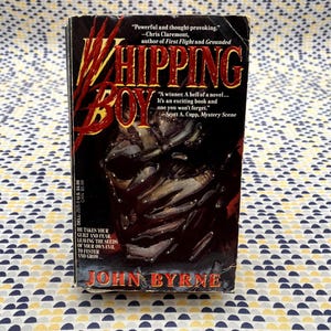 Op de afbeelding: Een paperback boek genaamd "Whipping Boy" van John Byrne. De omslag toont een donkere, getextureerde afbeelding met de titel in grote, rode en gele letters. Citaten van Chris Claremont en Scott A. Cupp zijn ook zichtbaar.