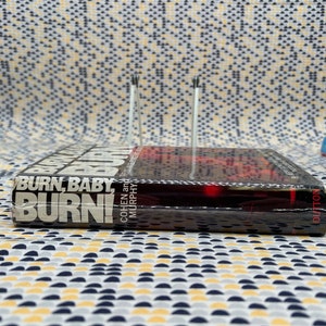 May include: A black hardcover book titled "Burn, Baby, Burn!" by Cohen and Murphy. The book is wrapped in plastic and has a red and black cover.
