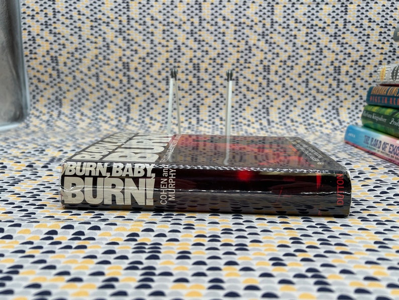 May include: A black hardcover book titled "Burn, Baby, Burn!" by Cohen and Murphy. The book is wrapped in plastic and has a red and black cover.